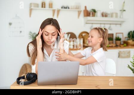 Une petite fille empêche sa mère de travailler de la maison. Travail à distance pendant la quarantaine. Une maman fatiguée s'assoit à un ordinateur portable, tenant sa tête. Banque D'Images