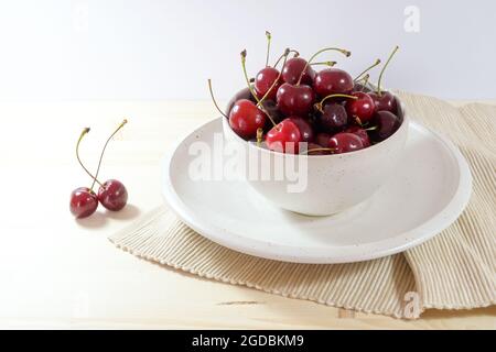 Cerises douces bio dans un bol en céramique blanc cassé et assiette avec une serviette sur une table en bois clair, fruits frais comme encas d'été, espace de copie, sélectionnez Banque D'Images