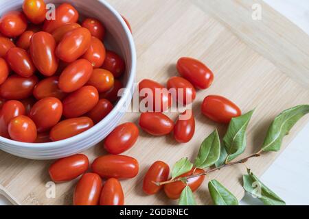 Bol de tomates cerises sur une planche à découper en bambou avec tomates cerises mûres rouges sur la planche à découper et branche de feuilles vertes séchées Banque D'Images