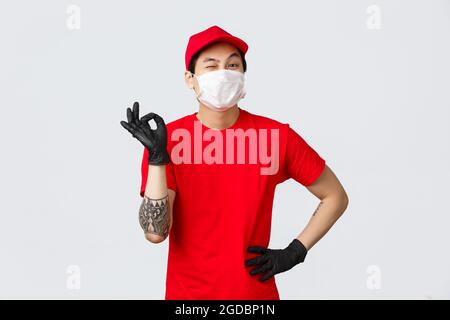 Pas de problème, livrer votre colis. Un bon préposé à la livraison asiatique en uniforme rouge, affiche et clin d'œil, assure la sécurité des emballages. Porter des gants de protection Banque D'Images