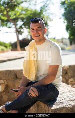 Un jeune homme heureux assis sur un banc de pierre avec un téléphone portable dans la main. Il est caucasien avec la peau brune. Il porte un T-shirt jaune et un tr noir Banque D'Images