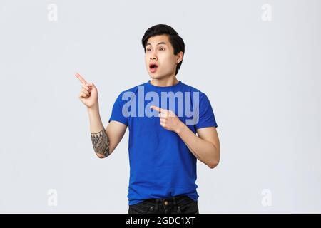 Choqué et sans voix, un homme asiatique qui voit quelque chose d'extraordinaire. Homme en t-shirt bleu avec tatouages, bouche ouverte et regardant dans le coin supérieur gauche stupéfié Banque D'Images