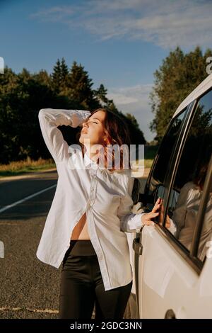 Une femme dans une chemise blanche à côté d'une voiture blanche sur la route. Un voyage dans la nature, repos en dehors de la ville. Photo verticale Banque D'Images