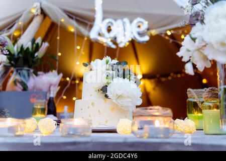 Gâteau de mariage à deux niveaux décoré de fleurs de pivoine fraîches et d'or comestible sur la table en bois avec fleurs, bougies. Mariage en extérieur avec lumières Banque D'Images