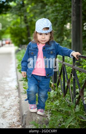 Petite fille caucasienne portant un Jean et un chapeau marchant le long d'une clôture en acier dans un parc Banque D'Images