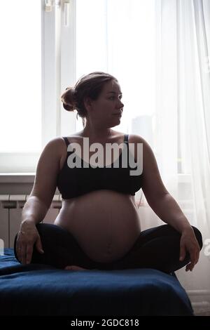 Belle femme enceinte assise au lit dans lotus poser avec les mains sur les genoux. Derniers mois de grossesse. Caucasien femelle Banque D'Images