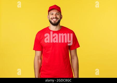 Livraison express du courrier, transfert de colis et concept logistique. Joyeux jeune barbu coursier dans le chapeau rouge uniforme et t-shrit, souriant heureux comme Banque D'Images