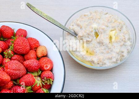 De délicieuses fraises mûres fraîches dans un bol en métal blanc et une assiette de porridge de flocons d'avoine avec beurre sur une table blanche. Banque D'Images