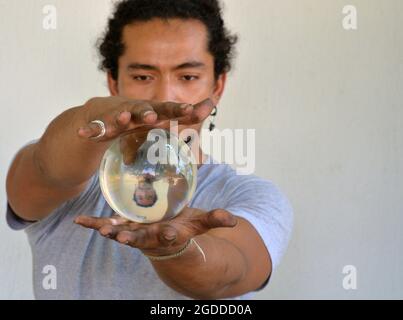 Beau jeune homme avec des ongles sales tient dans ses mains une boule de cristal avec sa propre image miroir retournée verticalement et regarde l'orbe. Banque D'Images