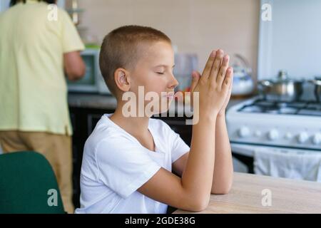 Un garçon caucasien prie à la maison à table avant de manger. Restez à la maison et priez à dieu. Culte en ligne de l'église le dimanche. Main de l'enfant pliée dans la prière. Fils assis dans la cuisine priant à dieu Banque D'Images