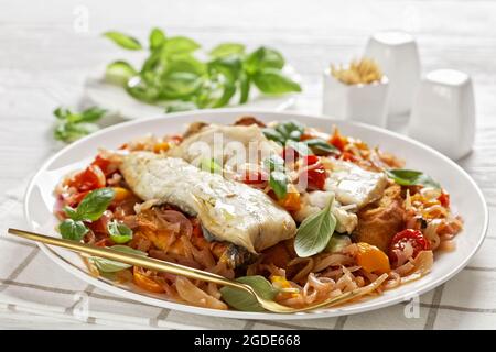 filet de poisson blanc poché dans une sauce à base de tomate et de vin blanc avec fenouil, herbes et pain crostini croustillant sur une assiette blanche, sur une table en bois Banque D'Images