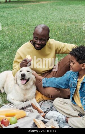 Heureux père assis sur l'herbe avec son fils pendant un pique-nique, ils jouent avec le chien Banque D'Images