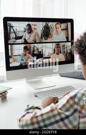 Vue arrière du petit garçon assis à la table devant le moniteur d'ordinateur et parlant à ses amis en ligne Banque D'Images