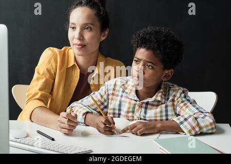 Petit garçon africain se concentrant sur ses devoirs il assis à la table avec la mère qui l'aide Banque D'Images