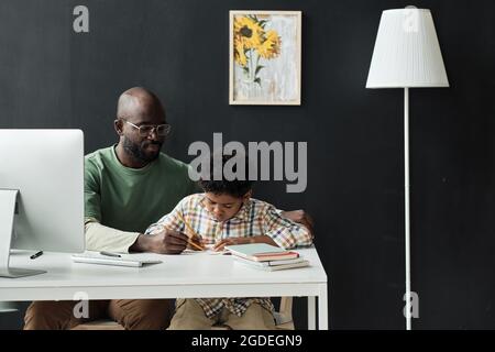 Un jeune enseignant africain enseignant à un petit garçon d'écrire pendant qu'ils sont assis à la table dans la salle de classe