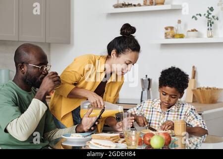 Une jeune femme verse du jus dans des verres pendant que sa famille prend le petit déjeuner à la table de la cuisine Banque D'Images