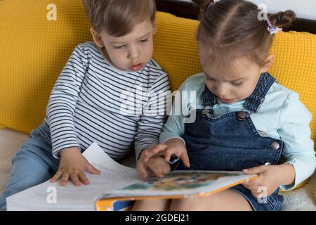 Deux enfants avant-garçonnets lisent un grand livre intéressant de contes de fées sur un lit jaune. Frères et sœurs petits jumeaux lecteur ont plaisir, heureux enfant sur Banque D'Images