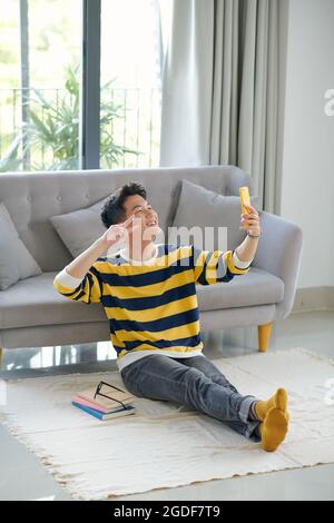 Un jeune homme s'assoit à la maison sur le sol dans le salon, prend des photos de lui-même sur l'appareil photo avant du téléphone et sourit Banque D'Images