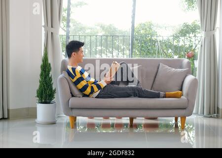 Portrait d'un jeune homme joyeux lisant le livre tout en étant assis sur un canapé dans son salon Banque D'Images