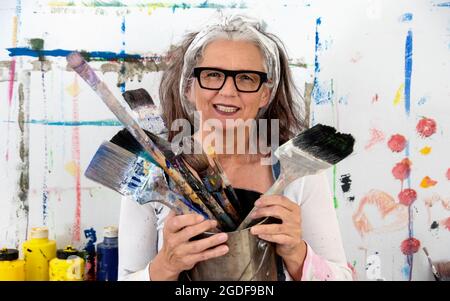 portrait chanceux et fier d'une femme mûre plus âgée souriante, artiste, dans ses années 50 avec des cheveux gris et des verres noirs, et beaucoup de grands pinceaux, espace de copie Banque D'Images