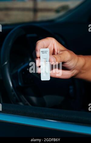 gros plan d'un jeune homme, assis dans le siège conducteur de sa voiture, tenant un dispositif d'essai covid-19 montrant un résultat négatif dans sa main Banque D'Images
