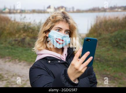 mignonne jeune femme, fille de l'adolescence dans le masque médical avec le sourire drôle dessiné. Prend des photos de selfie, communique en ligne. L'émotion de l'étonnement, de l'horreur. Grand rou Banque D'Images