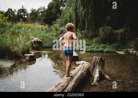 Vue arrière d'un garçon marchant pieds nus sur une bûche dans l'eau en cours d'eau en été Banque D'Images