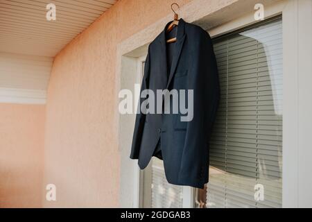 Une veste noire est suspendue contre une fenêtre blanche dans la pièce. Blazer classique classique bleu foncé brun noir pour hommes. Vêtements de mode pour hommes d'affaires Banque D'Images