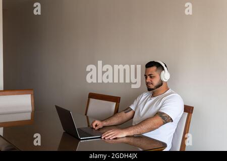 Latino homme avec des tatouages et des écouteurs allongé sur une chaise et souriant tout en regardant un ordinateur portable Banque D'Images