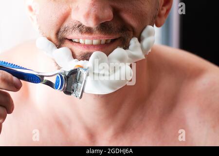 Beau sourire homme rasant sa barbe avec un rasoir dans la salle de bains. Procédure cosmétique quotidienne, concept de soins de la peau du visage Banque D'Images