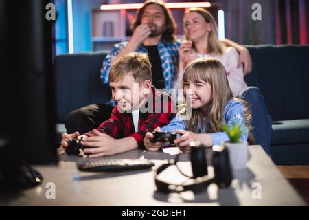 Mignon frère et soeur jouant des jeux vidéo avec des manettes tandis que les parents heureux se détendant sur le canapé avec pop-corn. Bonne famille caucasienne passer la soirée à la maison. Banque D'Images