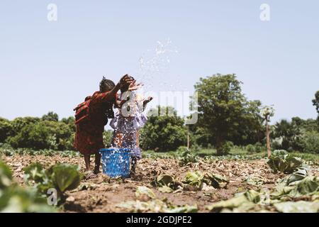 Deux filles dans les robes africaines traditionnelles debout sur la terre sèche sont l'eau étincelante notre d'un seau bleu Banque D'Images