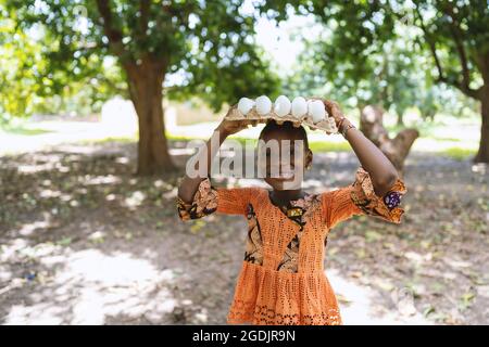 Jolie fille noire avec un plateau d'œufs blancs frais sur sa tête, souriant à l'appareil photo Banque D'Images