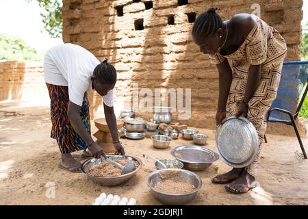 Dans cette image, deux jeunes femmes noires préparent un repas pour les membres de leur famille dans une cuisine en plein air dans un village ouest africain Banque D'Images