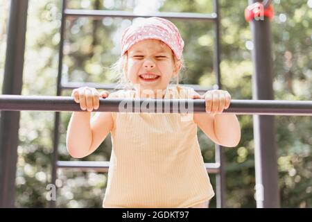 une petite fille se tire sur un bar horizontal dans un parc de la ville pendant une journée d'été Banque D'Images