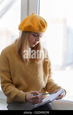 une jeune fille étudiante dans un béret jaune fait des croquis dans son carnet de croquis assis près de la fenêtre Banque D'Images