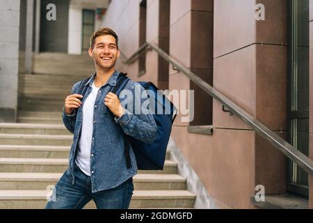 Photo moyenne de jeune homme joyeux et charmant de service de livraison de nourriture avec grand sac à dos thermique posé dans les escaliers du bâtiment de bureau Banque D'Images