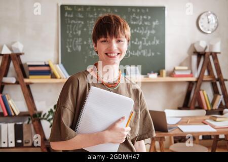 Portrait d'un jeune enseignant heureux avec des livres souriant à l'appareil photo tout en se tenant dans une salle de classe vide à l'école Banque D'Images