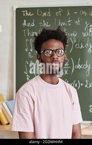 Portrait d'un étudiant africain en lunettes regardant l'appareil photo tout en se tenant près du tableau noir dans la salle de classe Banque D'Images