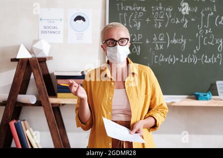 Portrait d'un jeune enseignant dans un masque de protection expliquant le nouveau matériel en étant debout dans la salle de classe Banque D'Images