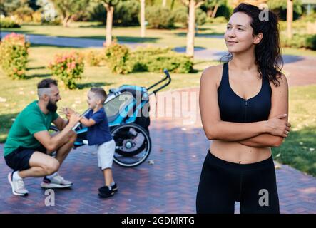 Portrait d'une maman sportive portant des vêtements de sport sur l'arrière-plan de son mari et de son fils après un entraînement et des exercices dans un parc de la ville. Bonne famille sportive Banque D'Images