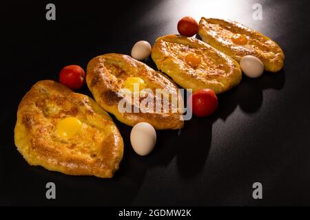 Khachapury adjarien cuit gros plan sur fond noir avec des tomates rouges et des œufs frais. Banque D'Images