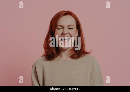 Une femme surjoyeuse aux cheveux rouges riant positivement, avec des yeux fermés, isolée sur fond rose Banque D'Images