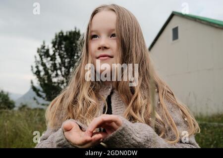 Petite fille avec de longs cheveux touchant des graines à la main et regardant loin avec le visage de rêve tout en passant du temps dans le champ près de la grange sur Hvalfjordur, Islande Banque D'Images