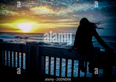 Jeune femme assise sur la clôture, regarde sur la mer au soleil couchant Banque D'Images