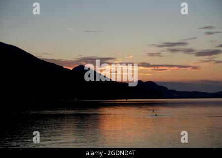 Un spectaculaire coucher de soleil d'orange se reflète dans l'eau cristalline du lac tandis que deux nageurs silhouettés profitent de la dernière baignade de la journée en face de la chaîne de montagnes. Banque D'Images