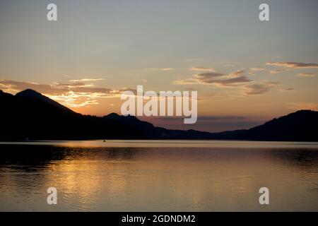 Coucher de soleil saisissant nuages illuminés suspendus avec élégance au-dessus de la chaîne montagneuse alpine se reflète dans l'eau étincelante translucide et rippée idyllique du lac. Banque D'Images
