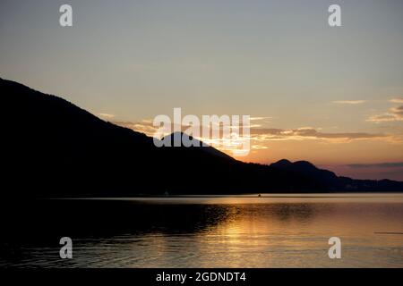 Coucher de soleil derrière une chaîne montagneuse alpine en pente avec des nuages jaunes bas suspendus et des reflets orange lumineux dans l'eau claire et ondulée du lac. Banque D'Images