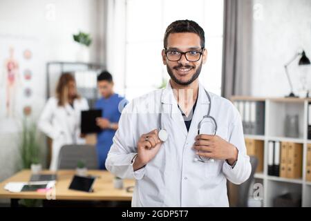 Un jeune homme sympathique, le médecin arabe malais, regarde la caméra et sourit tandis que ses collègues se tiennent debout et parlent en arrière-plan Banque D'Images