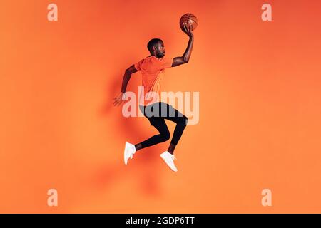 Athlète masculin sautant avec le ballon de basket. Vue latérale du sportif s'exerçant sur fond orange. Banque D'Images